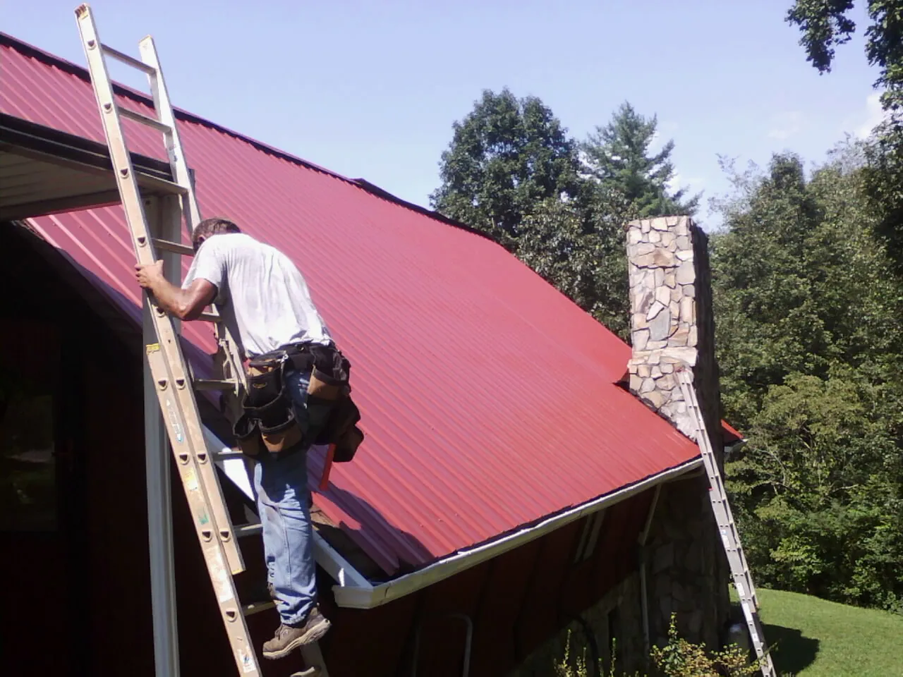 Roofer on a ladder beside a red metal roof and stone chimney — Soffit & Fascia Repair by Roofing Brothers in Dorset